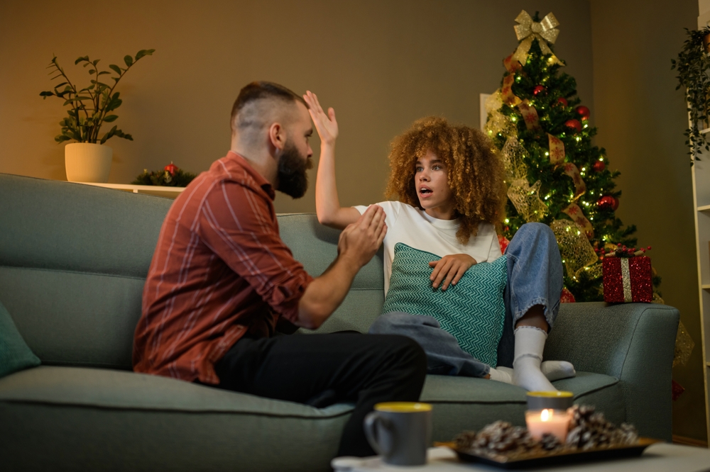 Young woman is sitting on a sofa with a shocked expression as a man gestures angrily at her